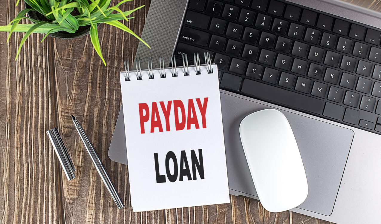 Notebook displaying the words “Payday Loan” placed beside a laptop, mouse, pen, and plant on a wooden desk, representing how payday loans work in Singapore.