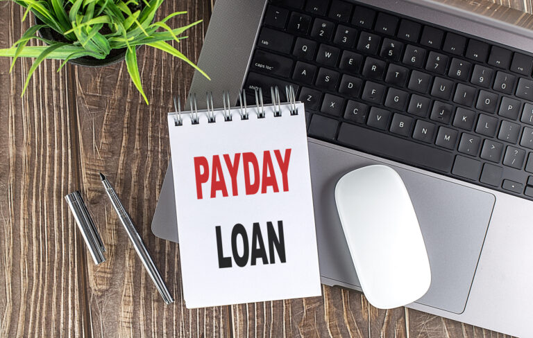 Notebook displaying the words “Payday Loan” placed beside a laptop, mouse, pen, and plant on a wooden desk, representing how payday loans work in Singapore.
